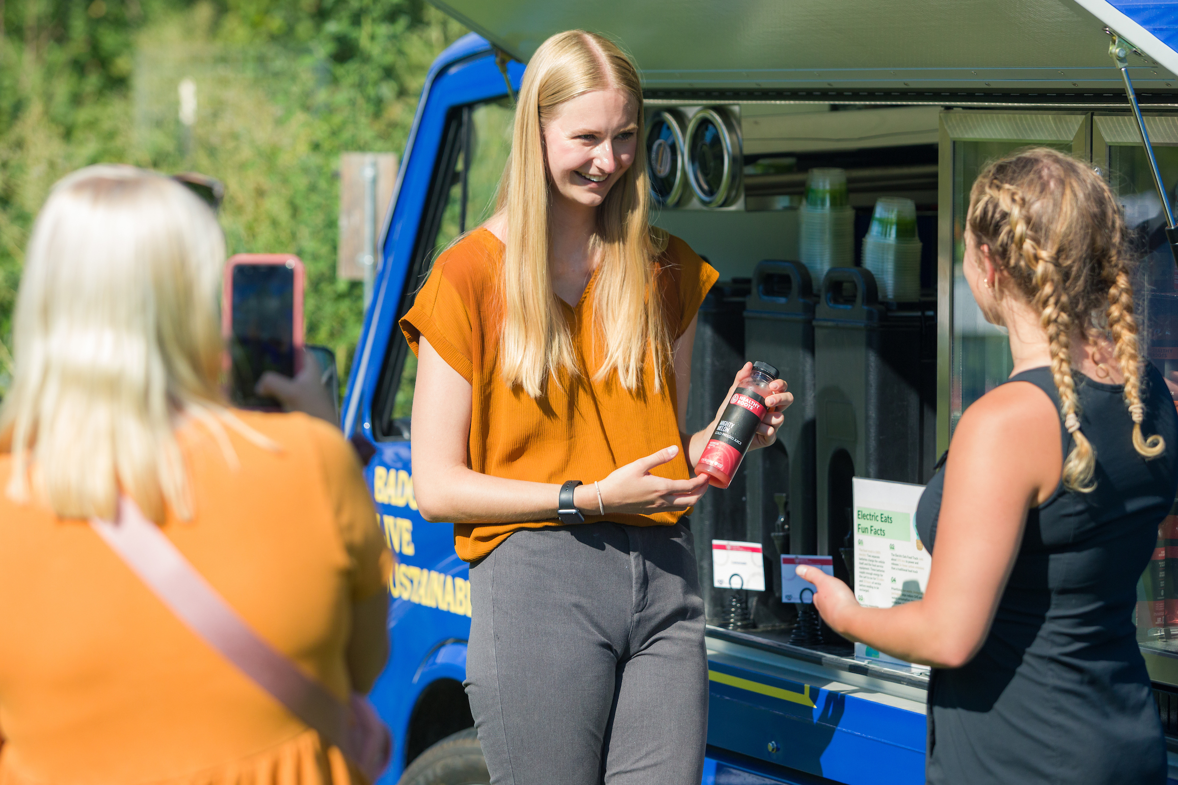 A student showing a beverage available at the Electric Eats food truck to another student while a third looks on 