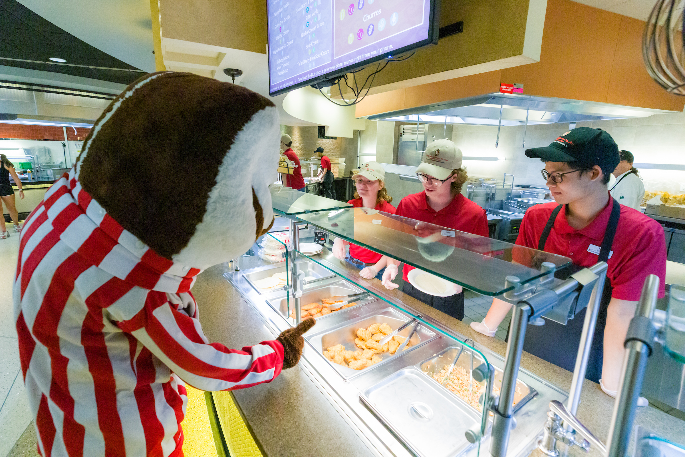 Bucky Badger selects food from the dining line while student staff look on